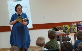 Na zdjęciu autorka mówi do mikrofonu, na stoliku książki, w tle dzieci siedzące na krzesłach.