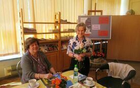 Dwie uśmiechnięte kobiety, jedna stoi i trzyma w ręku róże, druga siedzi.