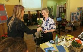 Kobieta w okularach i blond włosach wręcza kwiaty kobiecie w bluzce w kwiaty.