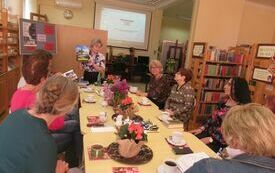 Grupa kobiet siedzących przy stoliku zwróconych w kierunku stojącej kobiety trzymającej w ręku książkę o Wielopolu Skrzyńskim.