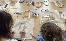 Kobieta odrysuje od szablonu portret kobiety na torbie ekologicznej. Obok inne kobiety odrysowują inne wzory.  