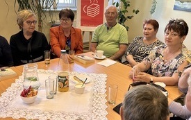 Grupa starszych ludzi siedzi dookoła stołu, w tle rośliny i plakat Dyskusyjnych Klubów Książki.
