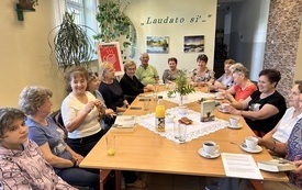 Grupa ludzi siedzi przy stole i rozmawia. w tle roślina, okno i plakat Dyskusyjnych Klubów Książki.