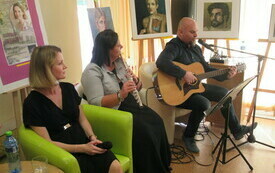 Mężczyzna śpiewa i gra na gitarze. Obok kobieta gra na flecie. Druga kobieta siedzi z mikrofonem w ręku.