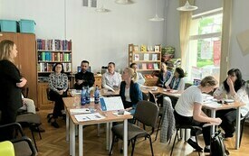 Uczestnicy spotkania siedzą przy stolikach zwróceni w kierunku kobiety w czarnej marynarce prowadzącej spotkanie.