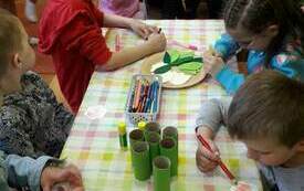 Grupa dzieci siedząca przy stolikach, na kt&oacute;rym znajdują się materiały plastyczne: kredki, kolorowe kulki, klej, papierowe rolki. 