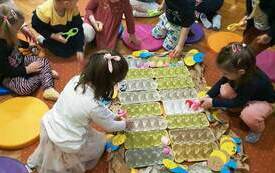 Grupa dzieci siedzi na dywanie wok&oacute;ł ułożonych obok siebie pudelek do przechowywania jaj. Dzieci układają w wytłaczankach kolorowe kulki. 