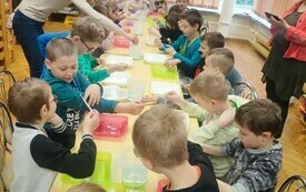Dzieci siedzą wok&oacute;ł stołu i wykonują doświadczenie z użyciem plastikowych pojemnik&oacute;w wody i pipet. 
