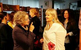 Kobieta w marynarce okularach i kręconych włosach rozmawia z kobietą w blond włosach, w białej bluzce z makiem i kolczykami w kształcie motyla. W tle inne kobiety.