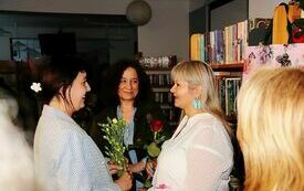 Kobieta w ciemnych włosach wręcza bukiet goździków gościowi spotkania, kobiecie w blond włosach. Obok stoi kobieta w ciemnych kręconych włosach.