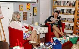 Dwie młode kobiety siedzą w fotelach przy stoliku kawowym, jedna trzyma w ręku otwartą książkę. Obok regały z książkami.