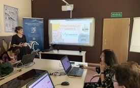Prowadząca szkolenie młoda kobieta w sukience w groszki i okularach stoi obok tablicy multimedialnej. Na tablicy prezentacja dotycząca komunikacji miejskiej. Przy stole siedzą uczestnicy szkolenia.