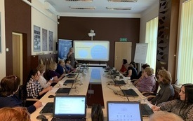 Grupa uczestników szkolenia siedząca przy stole przed laptopami. Na tablicy multimedialnej wyświetla się prezentacja. Obok stoi młoda kobieta prowadząca szkolenie.
