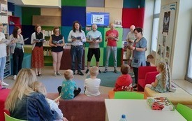 Osoby dorosłe czytają, dzieci stoją przed nimi i słuchają.