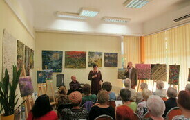 Publiczność zgromadzona na spotkaniu zwr&oacute;cona w kierunku stojącego mężczyzny i kobiety trzymających mikrofony. Na ścianach kolorowe gobeliny.