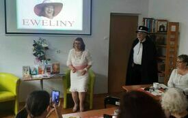 Kobieta w kremowej sukience stoi przed publicznością obok starszy mężczyzna z brodą w czarnej pelerynie i kapeluszu. 