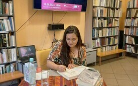 Sylwia Winnik podpisuje książki. W tle regały z książkami oraz na ekranie plakat Dyskusyjnych Klubów Książki.
