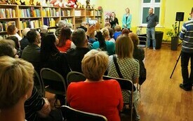 Grupa ludzi siedzi na krzesłach, kobieta, dyrektor biblioteki w Cieszanowie, wita gości.