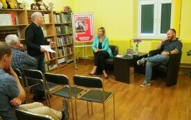 Na pierwszym planie pisarz Wojciech Chmielarz siedzi i rozmawia z bibliotekarką. Mężczyzna, uczestnik spotkania, stoi i mówi.