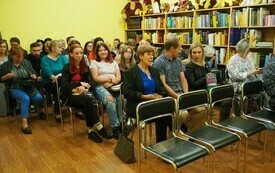 Grupa ludzi siedzi na krzesłach, z prawej strony regały z książkami.