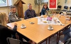 Grupa ludzi siedzi wokół stołu i rozmawia. W tle rośliny i plakat Dyskusyjnych Klubów Książki.