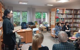 Grupa ludzi siedzi na krzesłach, jedna z kobiet stoi. Wokół regały z książkami.