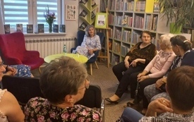 Kilkanaście osób siedzi w kole na krzesłach i słucha prowadzącej spotkanie.