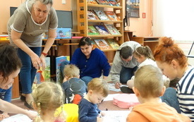Dzieci wraz z rodzicami siedzą na dywanie i rysują. Obok regał biblioteczny
