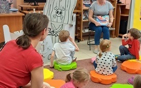 Grupa dzieci siedzi na dywanie, obok kobieta z książką na kolanach i statek piracki z papieru.