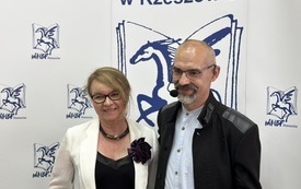 Mężczyzna z brodą w niebieskiej koszuli stoi obok kobiety w kremowej marynarce i czarnej bluzce. W tle ścianka z logo biblioteki: pegazem.