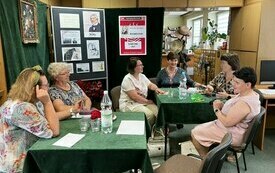Sześć kobiet siedzi przy nakrytych stolikach, w tle plakat Dyskusyjnych Klubów Książki i regały z książkami.