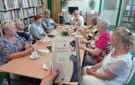 Na pierwszym planie dłoń trzyma książkę, w tle grupa kobiet siedzących przy stole. 