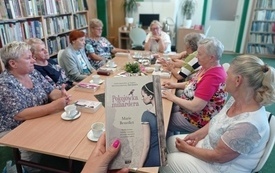 Na pierwszym planie dłoń trzyma książkę, w tle grupa kobiet siedzących przy stole.