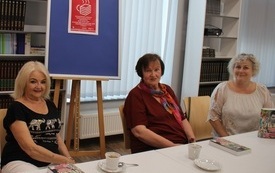 Trzy kobiety siedzą przy stole, w tle plansza z plakatem Dyskusyjnych Klubów Książki