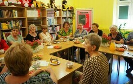 Grupa ludzi siedzi wokół stołu i rozmawia, na stole bukiety kwiatów, serwetki, pokrojony arbuz. Po lewej stronie regały z książkami.