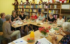 Grupa ludzi siedzi przy stole ustawionym w kształcie litery L. Na stole bukiety kwiatów, serwetki. W tle regały z książkami, a na regałach pluszowe misie różnej wielkości.