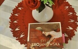 Na stole książka "Żona nazisty" Sylwii Trojanowskiej, obok wazon z bukietem róż.