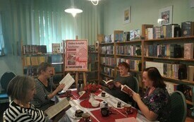 Cztery kobiety siedzą przy stole i trzymają w rękach książkę, czytają. W tle plakat Dyskusyjnych Klubów Książki.