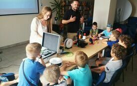 Mężczyzna i kobieta prezentują drukarkę 3D dzieciom. Przy stoliku dzieci oglądają figurki wydrukowane metodą 3D.