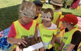 Dzieci w odblaskowych kamizelkach, z maskami na oczach, odgadują co znajduje się w pudełku. 