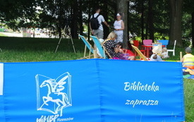 Na niebieskim parawanie napis Biblioteka zaprasza i logo z pegazem. W tle pod drzewami grupa os&oacute;b zgromadzonych na spotkaniu