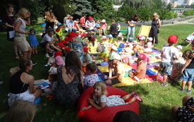 Uczestnicy spotkania plenerowego - dzieci wraz z opiekunami siedzą na kolorowych matach, pufach i leżakach. 