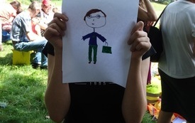 Dziecko w kapeluszu trzyma w rękach rysunek z postacią w kr&oacute;tkich włosach, zielonych spodniach i granatowej bluzce