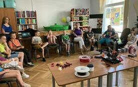 Dzieci uczestniczące w spotkaniu zgromadzone wok&oacute;ł stolika, na kt&oacute;rym stoi urządzenie do druku 3D.