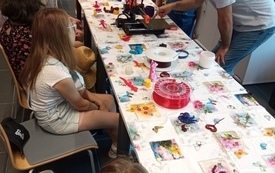 Grupa dzieci siedzi przy stoliku w bibliotece. Na stoliku drukarka 3D oraz figurki wydrukowane przy użyciu tej drukarki.
