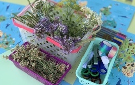 Na stoliku kolorowe kartki, flamastry, zioła, kwiaty, wstążki, nożyczki i kleje. 
