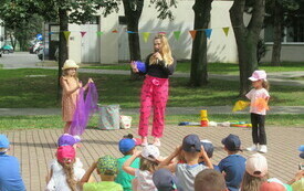 Dwie dziewczynki trzymają w rękach kolorowe chusty. Obok kobieta w r&oacute;żowych spodniach. Przed nimi grupa dzieci. 