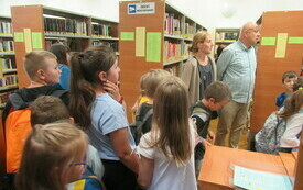 Grupa dzieci pomiędzy regałami bibliotecznymi. 