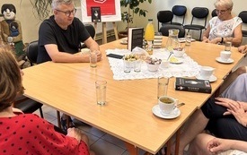 Dookoła stołu siedzi grupa ludzi, na stole filiżanki, książki, w tle rośliny i plakat Dyskusyjnych Klubów Książki