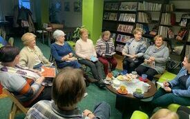 Grupa ludzi siedzi wokół stolika, na którym znajdują się kupki. W tle regały z książkami.